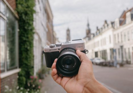 OM SYSTEM PEN Kamera mit Objektiv in einer Hand vor einer Häuserzeile